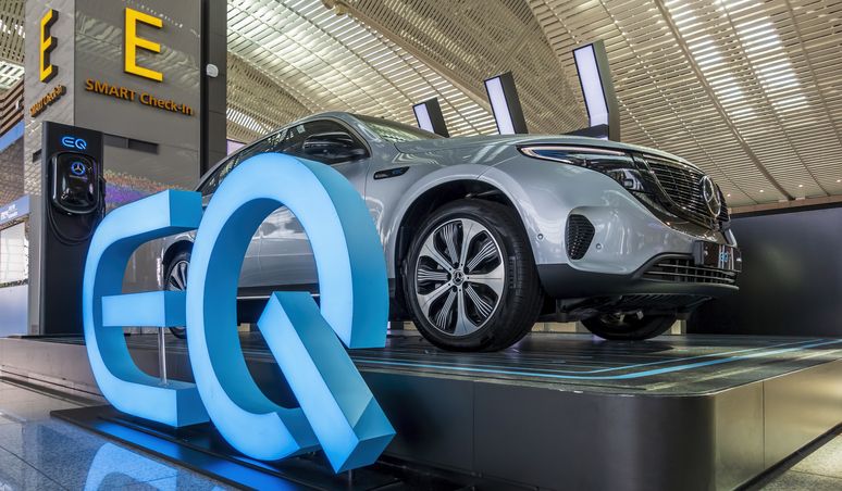 Silberner Elektro-SUV, ausgestellt auf einer Automobilausstellung, mit großem beleuchteten „EQ”-Schriftzug an der Front, unter einer modernen, hell beleuchteten Decke.