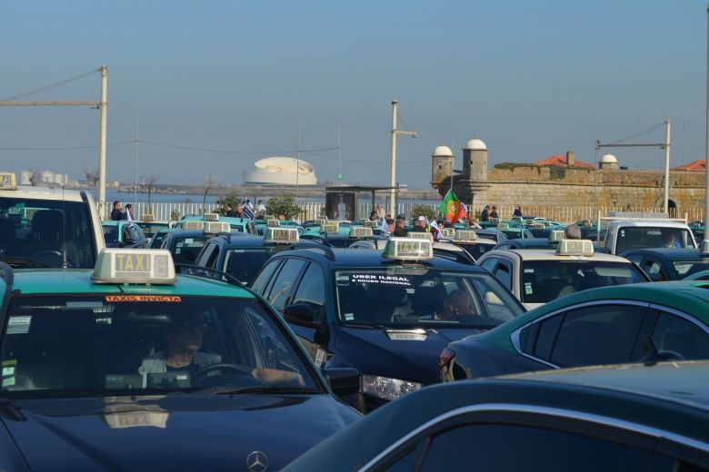 A large group of taxis is parked closely together near a waterfront, with drivers visible inside. A few people and flags are in the background.