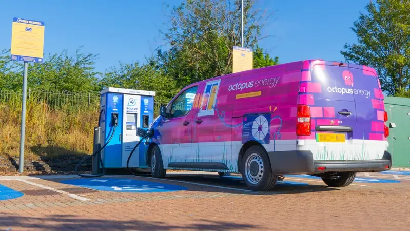A pink Octopus Energy van charging at an electric vehicle station in a sunny, tree-lined parking area.