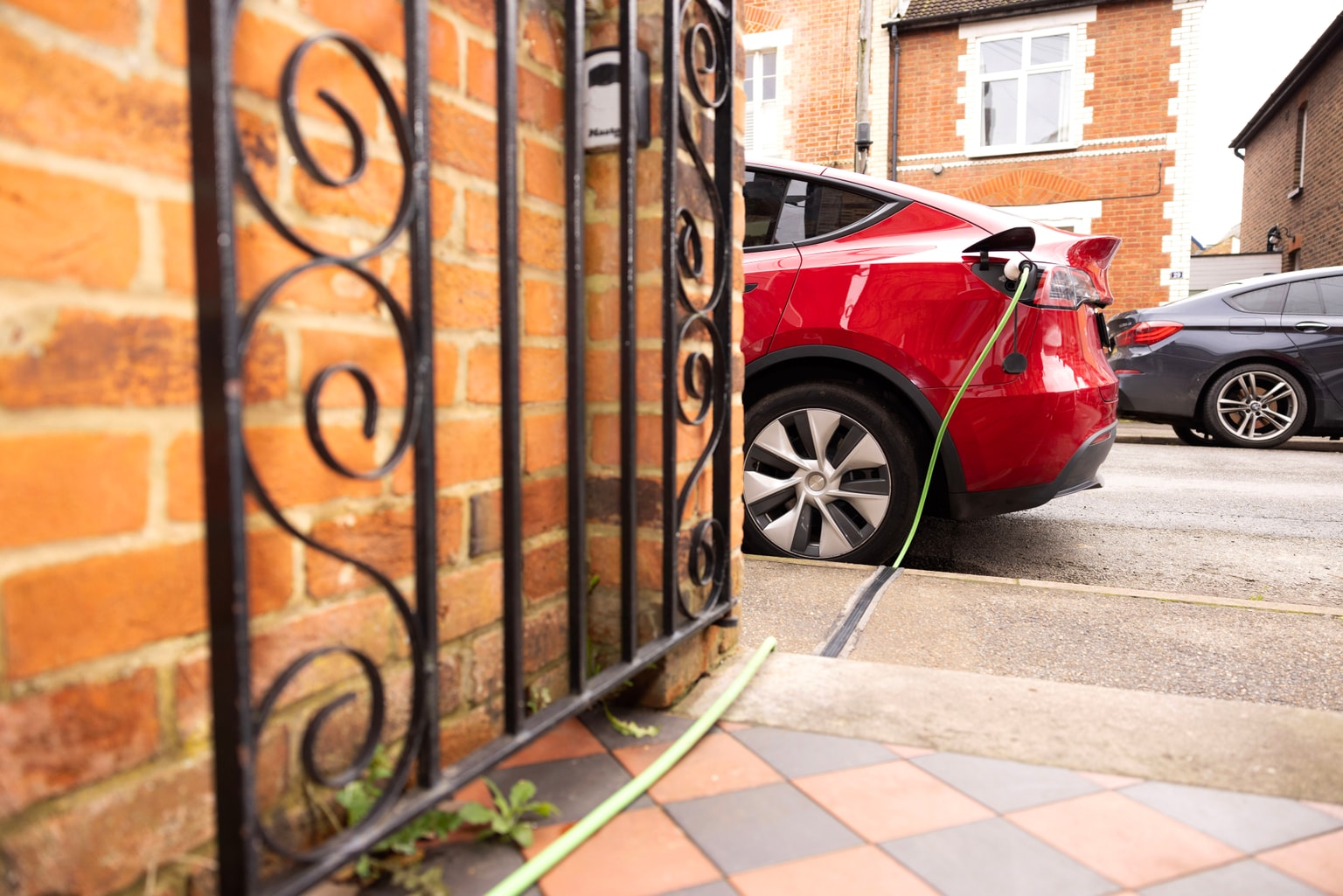 Coche eléctrico rojo cargándose en una calle residencial, conectado a un cable a través de una verja, con edificios de ladrillo al fondo.