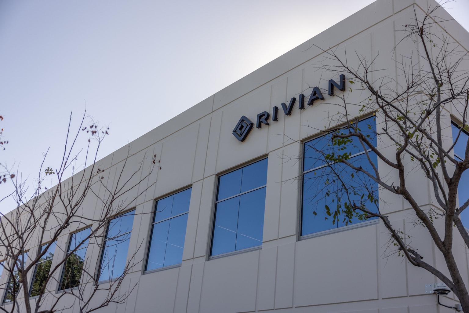 Vista exterior de un edificio con el logotipo de «Rivian», con grandes ventanas y árboles sin hojas en primer plano.