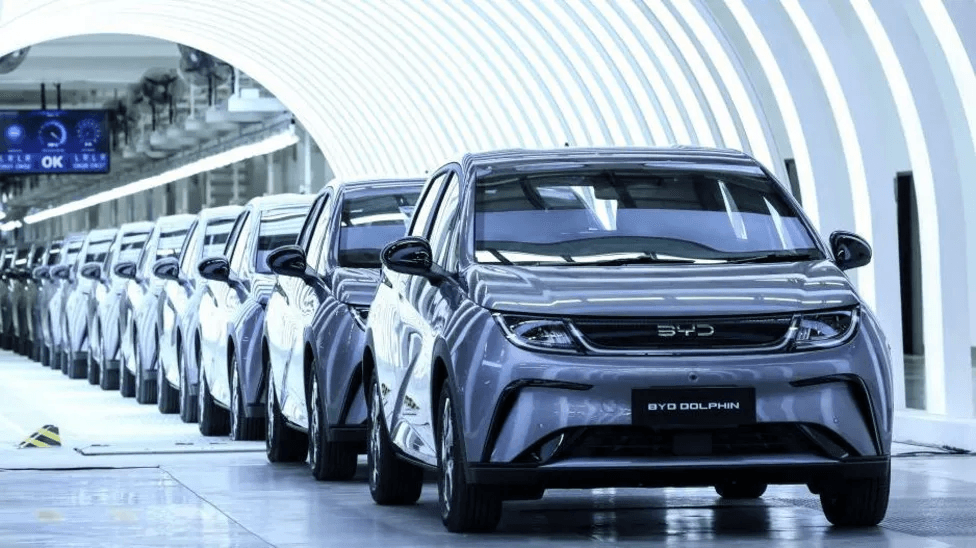 Una fila de automóviles BYD Dolphin plateados alineados en una línea de producción de una fábrica automotriz moderna y bien iluminada.